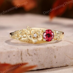 May include: A gold ring with a floral design and a central red gemstone. The ring has embossed flowers and leaves, with a small diamond accenting one of the flowers. The ring is set on a neutral surface.
