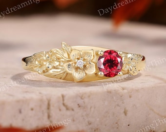 May include: A gold-colored ring with floral detailing and a central red gemstone. The ring features embossed flowers and leaves, with a small diamond accenting one of the flowers. The ring is set on a neutral surface.