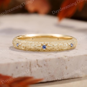 以下が含まれることがあります： 繊細な花柄と3つの小さな青い宝石が特徴のゴールドリング。リングはニュートラルな背景に配置され、その詳細なデザインと職人技を強調しています。リングの表面には繊細な花柄が刻まれています。
