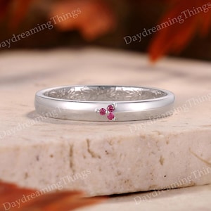 May include: A silver-toned ring featuring a smooth, polished band with a delicate floral engraving on the inner surface. The exterior of the band is accented with three small, round, vibrant pink stones arranged in a cluster on the front side.
