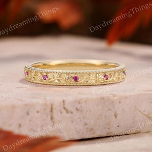 Op de afbeelding: Een gouden ring met een ingegraveerd bladpatroon op het buitenoppervlak. De ring is versierd met kleine, ronde, roze edelstenen die met tussenpozen zijn geplaatst. De randen van de ring zijn afgewerkt met een fijne, kralenachtige touwstructuur.