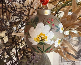 May include: A hand-painted Christmas ornament with a matte finish. The ornament features white flowers with yellow centers and red berries on a muted brown background. It hangs from a red velvet ribbon.