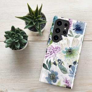 May include: A white smartphone case with a watercolor floral design featuring blue, purple, and green flowers. The case has a camera cutout and is next to two small potted succulents.