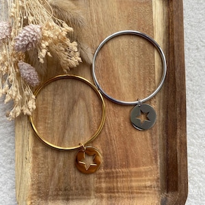 May include: Two circular bangles, one gold-toned and one silver-toned, each featuring a dangling circular charm with a star-shaped cutout. The bracelets are displayed alongside a cluster of dried, light-coloured, feathery botanical stems.