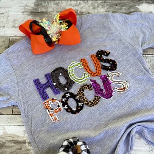 May include: A gray t-shirt with the words "Hocus Pocus" spelled out in colorful fabric patches. The patches are decorated with Halloween-themed patterns, including pumpkins, bats, and ghosts.