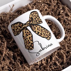 Op de afbeelding: Witte keramische mok met een luipaardprint strik ontwerp. De mok heeft een gebogen handvat en de naam "Sabrina" in cursief. De mok zit in een witte doos met bruine papieren vulling.
