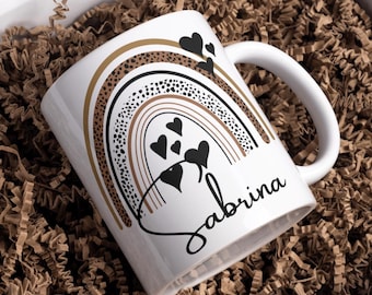 May include: White ceramic mug with a rainbow design in gold, brown, and black, adorned with black hearts. The name "Sabrina" is written in a cursive font. The mug is presented in a white gift box with brown paper filler.