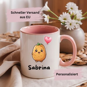 Può includere: Tazza in ceramica bianca con bordo e manico rosa. La tazza presenta una patata di cartone animato che tiene un palloncino a forma di cuore rosa. Il testo "Sabrina" è stampato sotto la patata. Il testo "Personalisiert" è stampato nell'angolo in basso a destra della tazza.