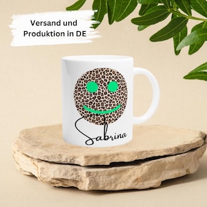 Può includere: Tazza in ceramica bianca con un'emoticon a stampa leopardata e il nome Sabrina scritto in corsivo nero. La tazza ha un contorno verde.