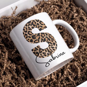 May include: White ceramic mug with a leopard print letter "S" and the name "Sabrina" written in cursive.