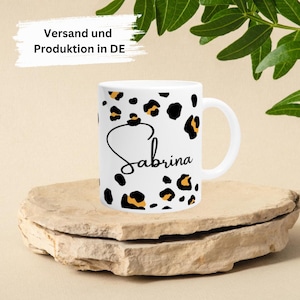 Peut inclure: Une tasse en céramique blanche avec un motif léopard noir et orange. Le nom "Sabrina" est écrit en cursive noire sur la tasse.