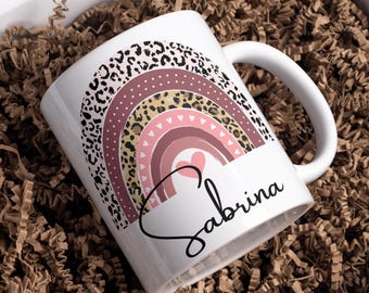 May include: White ceramic mug featuring a colorful rainbow design with leopard print and hearts. The name "Sabrina" is written in a cursive font. The mug is presented in a white box with brown paper filler.