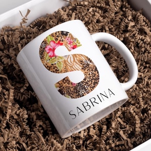 May include: White ceramic mug with the name "SABRINA" printed in black. The letter "S" is decorated with a floral and animal print design. The mug is presented in a white box with brown paper filler.