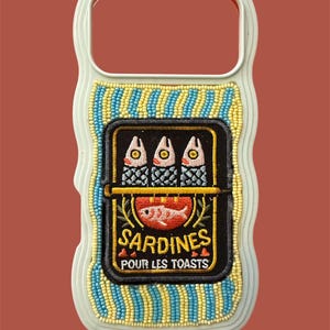 May include: A white phone case featuring a rectangular embroidered patch of three sardines in a tin. The patch includes the text SARDINES POUR LES TOASTS and is surrounded by vertical stripes of light blue and yellow seed beads on a white background.