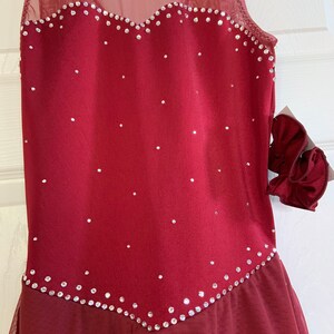 May include: Burgundy sleeveless figure skating dress with a mesh overlay and a heart-shaped neckline. The dress is decorated with rhinestones.