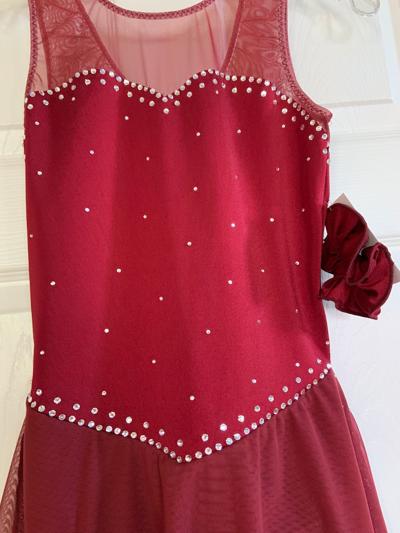 May include: Burgundy sleeveless figure skating dress with a mesh overlay and a heart-shaped neckline. The dress is decorated with rhinestones.