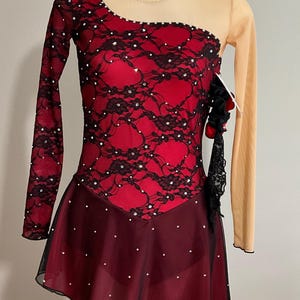 Puede incluir: Un vestido de patinaje artístico rojo y negro con una superposición de encaje y pedrería. El vestido tiene mangas largas y una falda con un dobladillo festoneado.