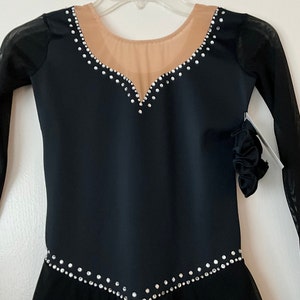 Op de afbeelding: Zwarte kunstschaatsjurk met lange mouwen en een transparante mesh-overlay. De jurk heeft een V-hals en is versierd met strass.