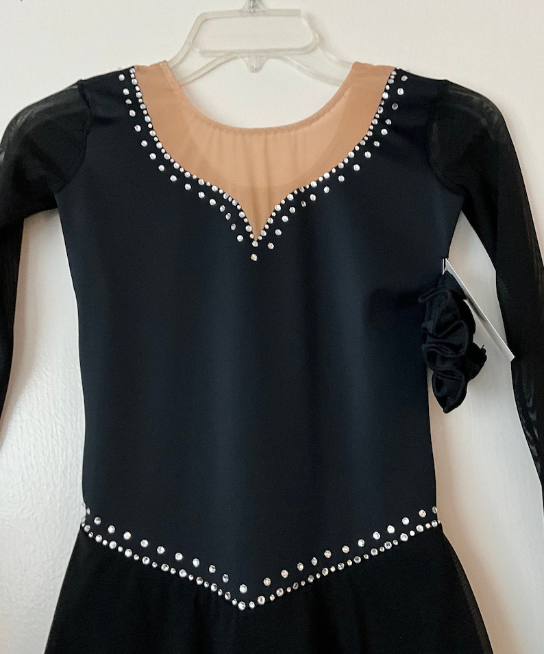 Op de afbeelding: Zwarte kunstschaatsjurk met lange mouwen en een transparante mesh-overlay. De jurk heeft een V-hals en is versierd met strass.