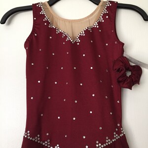 Puede incluir: Vestido de patinaje sobre hielo de color burdeos sin mangas, con un escote de malla transparente y una falda con un dobladillo festoneado. El vestido est&aacute; decorado con pedrer&iacute;a plateada.