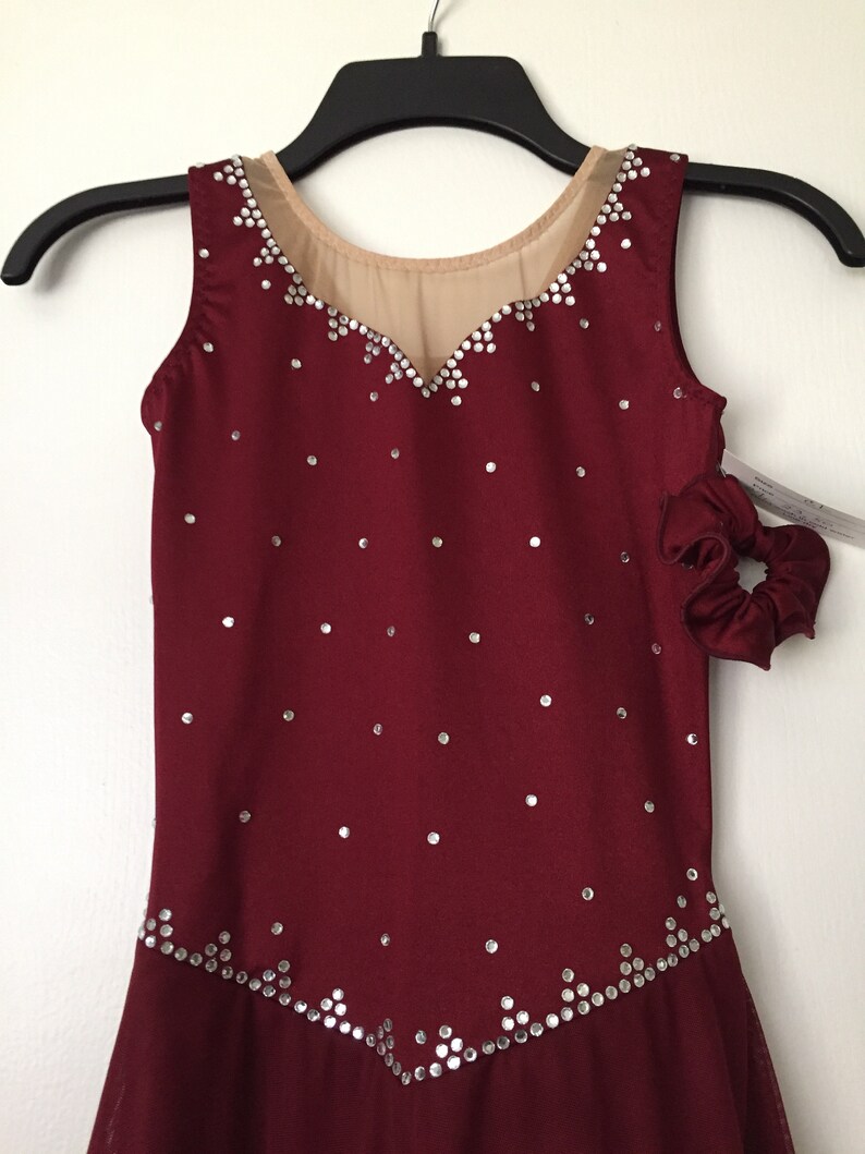 Puede incluir: Vestido de patinaje sobre hielo de color burdeos sin mangas, con un escote de malla transparente y una falda con un dobladillo festoneado. El vestido est&aacute; decorado con pedrer&iacute;a plateada.