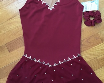 Puede incluir: Vestido de patinaje artístico de color burdeos con un escote de malla transparente y una falda con borde festoneado y pedrería. El vestido tiene un diseño simple y elegante.