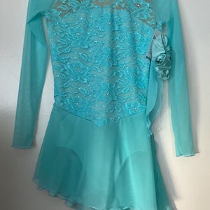 May include: A long-sleeved turquoise ice skating dress featuring a sheer bodice with floral lace overlay and scattered rhinestones. The skirt is made of layered chiffon with an asymmetrical hemline and a decorative fabric flower on the side.