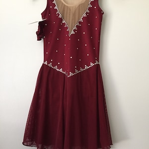 Puede incluir: Vestido de patinaje art&iacute;stico de color burdeos con un panel de malla transparente y un escote en V. El vestido est&aacute; decorado con diamantes de imitaci&oacute;n.