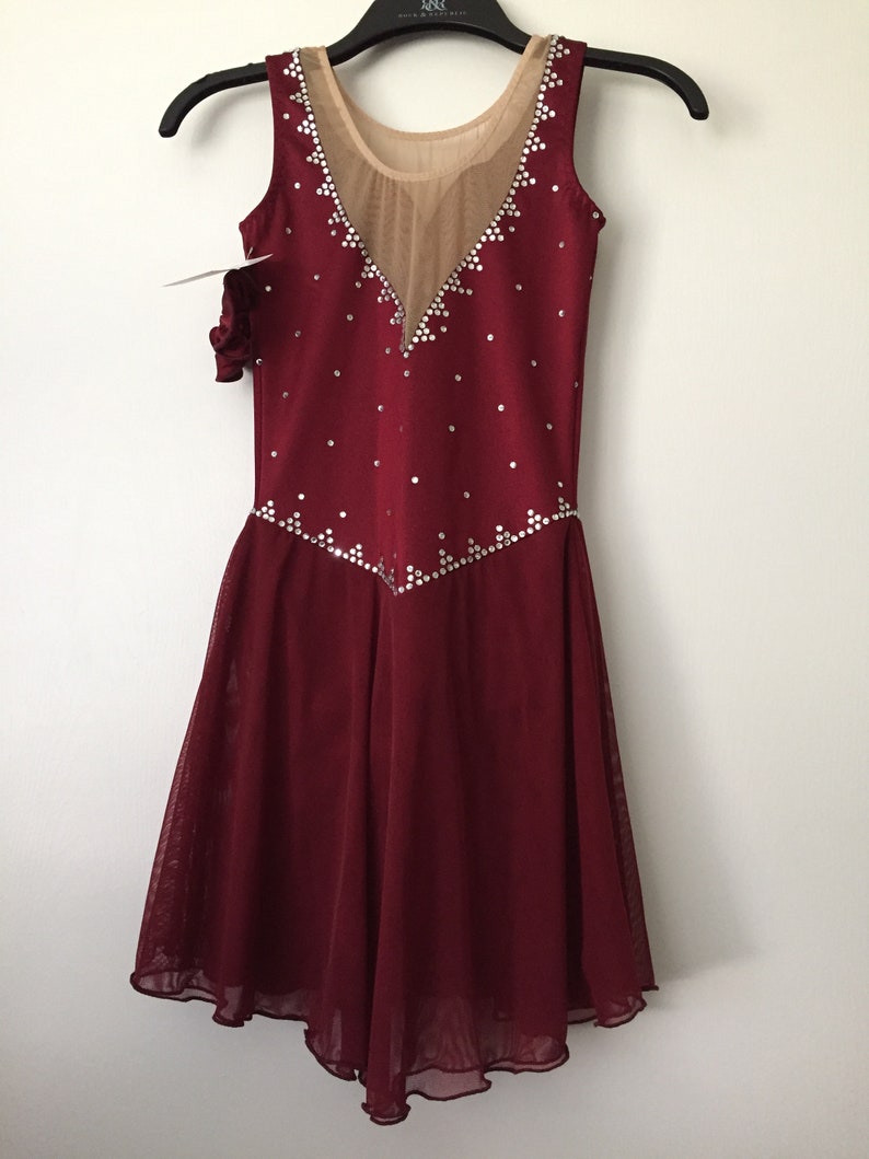 Puede incluir: Vestido de patinaje art&iacute;stico de color burdeos con un panel de malla transparente y un escote en V. El vestido est&aacute; decorado con diamantes de imitaci&oacute;n.
