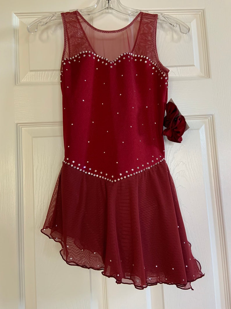 May include: Burgundy figure skating dress with a sheer mesh overlay and a heart-shaped neckline. The dress is decorated with rhinestones.