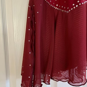 May include: A burgundy skating dress with a sheer overlay and a scalloped hem. The dress is decorated with rhinestones.