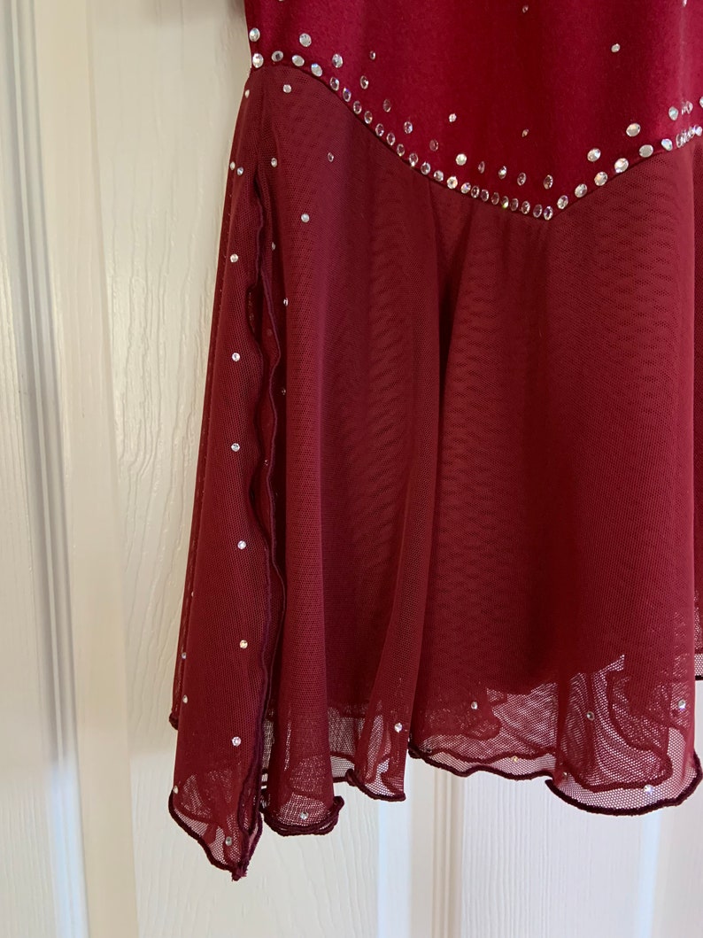 May include: A burgundy skating dress with a sheer overlay and a scalloped hem. The dress is decorated with rhinestones.