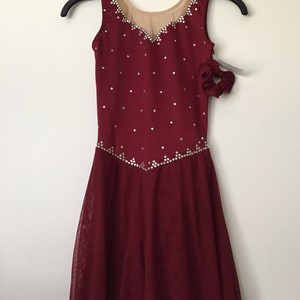 Puede incluir: Vestido de patinaje art&iacute;stico de color burdeos con un corpi&ntilde;o transparente y una falda con un dobladillo festoneado. El vestido est&aacute; decorado con pedrer&iacute;a plateada.