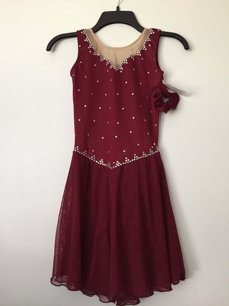 Puede incluir: Vestido de patinaje art&iacute;stico de color burdeos con un corpi&ntilde;o transparente y una falda con un dobladillo festoneado. El vestido est&aacute; decorado con pedrer&iacute;a plateada.