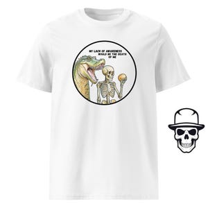 May include: White t-shirt with a black circle graphic featuring a cartoon alligator and skeleton. The text inside the circle reads "My lack of awareness would be the death of me."