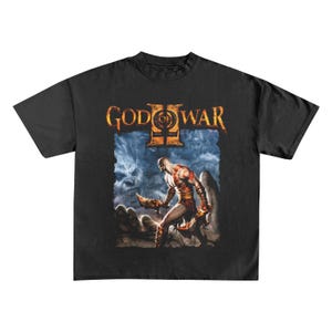 May include: Black t-shirt with the "God of War II" logo and a graphic of a warrior with blades. The design features a blue and orange colour scheme, with a detailed illustration of a character in a battle scene.