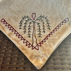 May include: Beige square fabric with a hand-stitched design. A weeping willow tree is embroidered in brown and green thread, with a heart at the top. The edges are trimmed with a red zig-zag stitch.