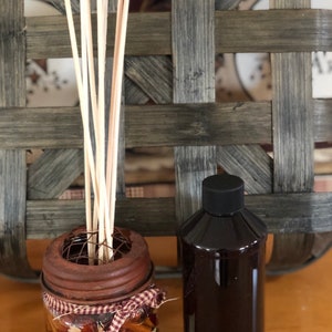 May include: A decorative reed diffuser set with a rustic aesthetic. The set includes a glass jar filled with dried fruit and spices, a separate bottle of dark liquid, and wooden reeds. The jar has a rusty metal lid and is tied with a plaid ribbon.