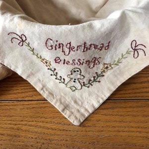 May include: Cream-colored, embroidered fabric with the words "Gingerbread Blessings" in burgundy thread. The design includes a gingerbread man and floral accents in green and gold. The fabric is folded over a wooden surface.