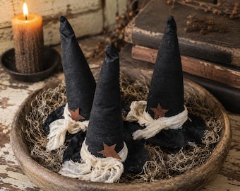 May include: Three black fabric witch hats with pointed crowns, each featuring a small rusty brown star accent and a frayed off-white cheesecloth band around the base, arranged inside a large, round, weathered wooden bowl filled with dried moss.