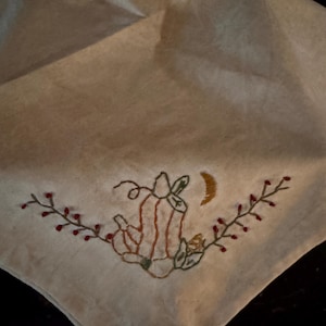 May include: Off-white, square-shaped cloth with an embroidered pumpkin and crescent moon design. The pumpkin is orange and brown, with green leaves and a brown stem. Red berries adorn the vines.