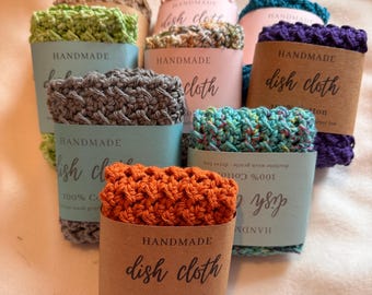 May include: A collection of handmade dish cloths in various colors, including orange, green, gray, pink, and blue. Each cloth is wrapped in a paper band that reads "HANDMADE dish cloth" and "100% Cotton". The cloths are arranged on a soft, white surface.