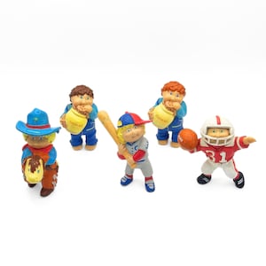 Peut inclure: Cinq figurines Cabbage Patch Kids, chacune vêtue d'une tenue différente. L'une porte un chapeau de cowboy et tient un cheval, une autre porte un uniforme de baseball et tient une batte, une autre porte un uniforme de football et tient un ballon de football, et les deux autres portent des tenues bleues et tiennent des biscuits.