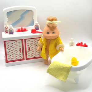 May include: A Cabbage Patch Kids doll with blonde hair in a yellow robe stands next to a white bathtub with a yellow rubber duck and a yellow towel. Beside it is a white vanity with a mirror, red accents, and bottles labelled CPK Baby.