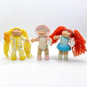 May include: Three vintage Cabbage Patch Kids dolls. One doll has yellow hair and a yellow teddy bear. Another doll wears a pink bonnet and a light pink outfit. The third doll has red pigtails and a blue dress.