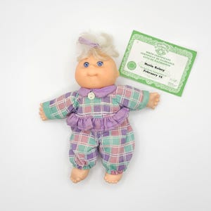 Peut inclure: Poupée Cabbage Patch Kids vintage, Marin Kelsey, vêtue d'une tenue à carreaux violet, vert et rose. La poupée a des cheveux blonds, des yeux bleus et un certificat de naissance. La tenue comprend un col à volants et un pantalon assorti.