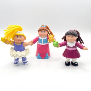Peut inclure: Trois poupées vintage Cabbage Patch Kids. Une poupée porte un justaucorps bleu et un tutu blanc, tenant une étoile. Une autre poupée porte une robe rose et tient un ours en peluche et un cadeau vert. La troisième poupée porte une robe bordeaux et tient un cadeau vert.