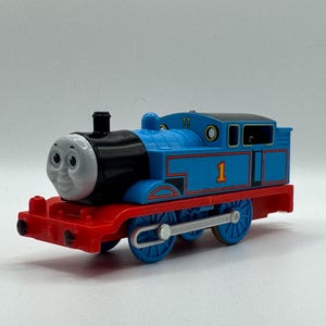 May include: A blue toy train with a smiling face, black smokestack, and red detailing. The number 1 is visible on the side. The train has blue wheels and a red base.