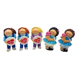 May include: Five Cabbage Patch Kids dolls, each with a different hairstyle and outfit. The dolls are holding hearts that say "I Love You" and ice cream cones.