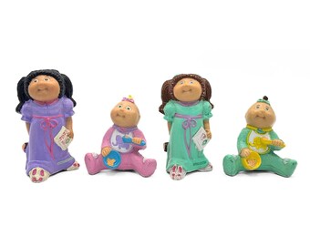 May include: Set of four Cabbage Patch Kids figurines. The figurines are dressed in pajamas and holding various accessories. One figurine is wearing a purple outfit, another is wearing a pink outfit, another is wearing a green outfit, and the last one is wearing a green outfit.